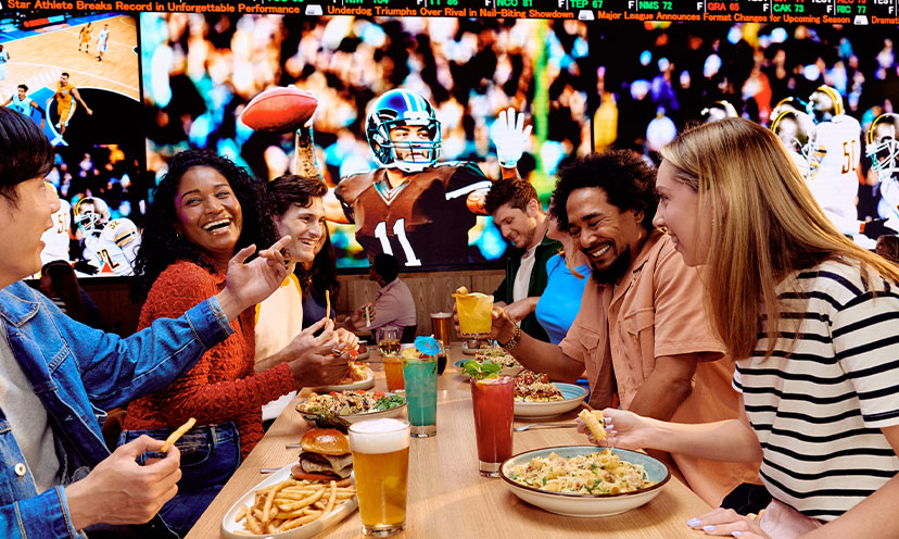 Image of several people at a restaurant table with food and drinks on it and a football game on a TV in the background